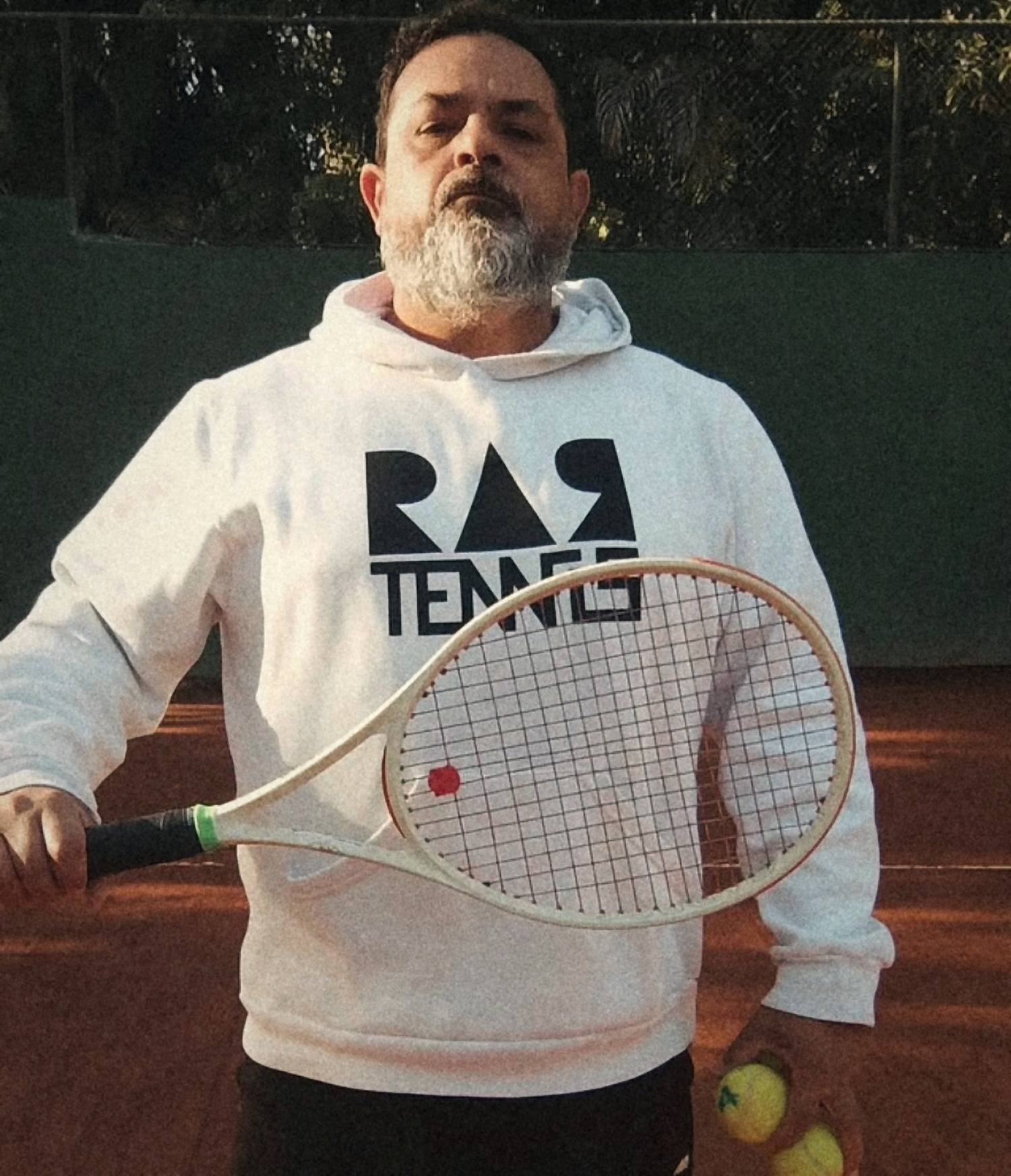 Rodrigo Aranha, professor da RAR Tennis, de moletom branco com o logo, segurando a raquete na quadra de saibro.
