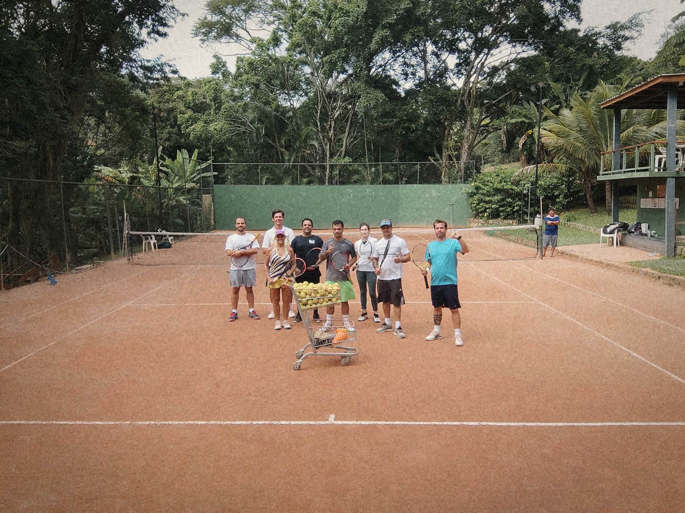 Turma de alunos da RAR Tennis de pé na quadra de saibro, com raquetes e o carrinho cheio de bolas, a mata e a varanda ao fundo.